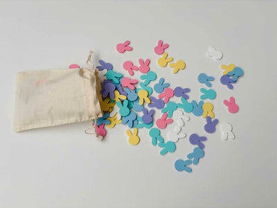 Ein kleiner, beiger Stoffbeutel liegt auf einer weißen Oberfläche. Daraus sind zahlreiche bunte, flache Hasen-Konfetti-Stücke in Pastellfarben – Rosa, Blau, Gelb, Lila und Weiß – herausgefallen. Die Hasenformen haben lange Ohren und verteilen sich spielerisch über die Fläche.