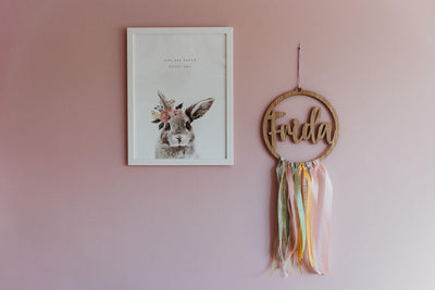 Namenskranz aus Holz mit Farbbändern Namens Frida hängend an einer pinken Wand neben ein Bild