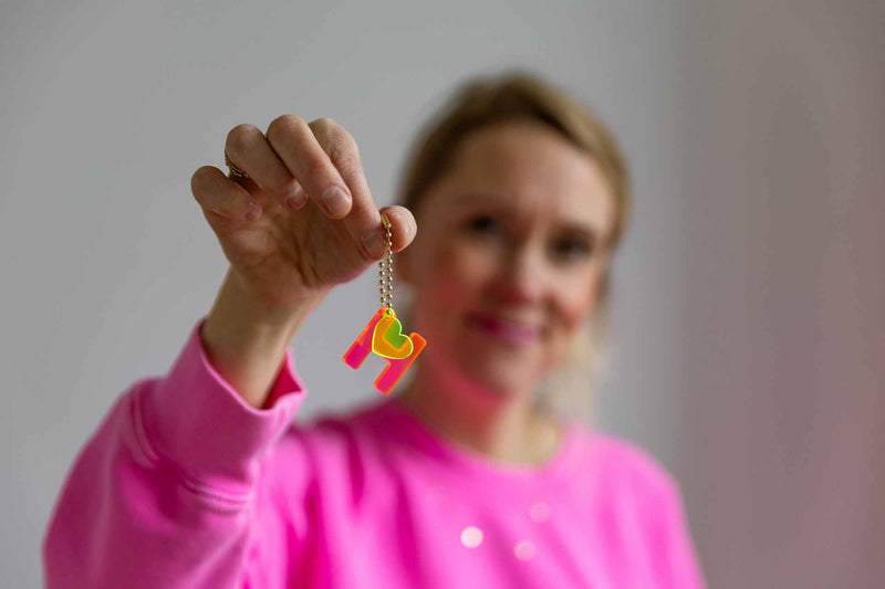 Eine Frau im Hintergrund hält einen Schlüsselanhänger aus Acryl mit einem neonpinken H und einem gelben Herz an einer goldenen Kette im Vordergrund