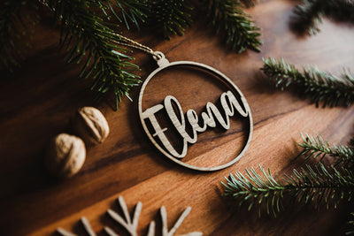 Weihnachtskugel aus Holz mit dem Namen Elena liegend neben Tannenzweigen. 